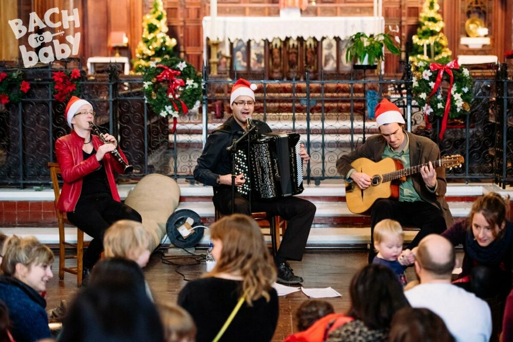 Three musicians wearing santa hats playing the clarinet, accordion, and guitar. Christmas trees behind them and an audience of parents and children in front