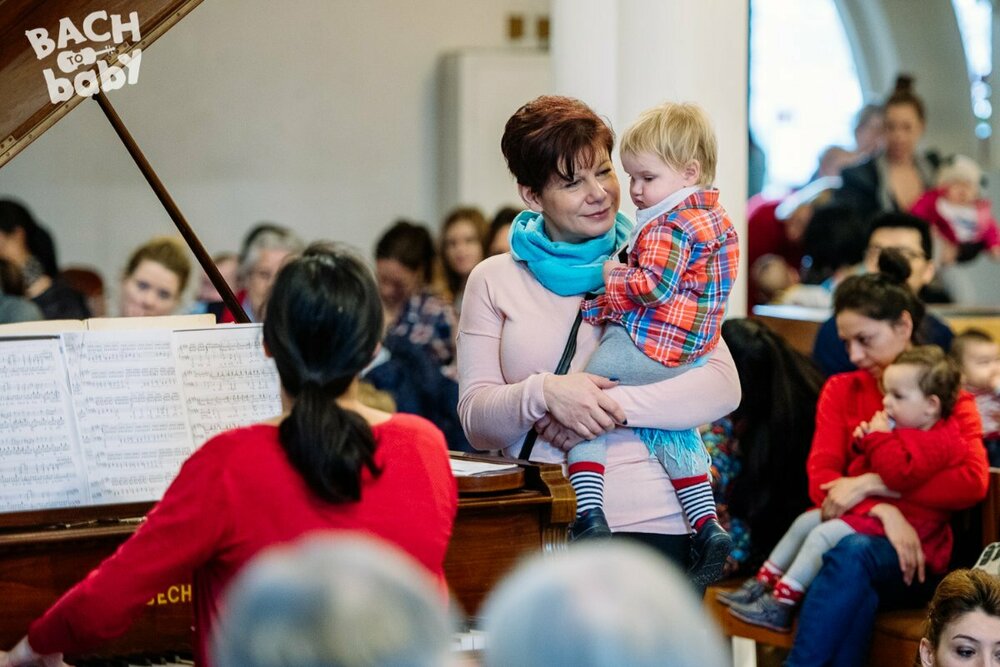 A pianist wearing a red top playing a grand piano. A lady with short red hair holding a young toddler stands next to her, and there are parents and babies watching around the piano