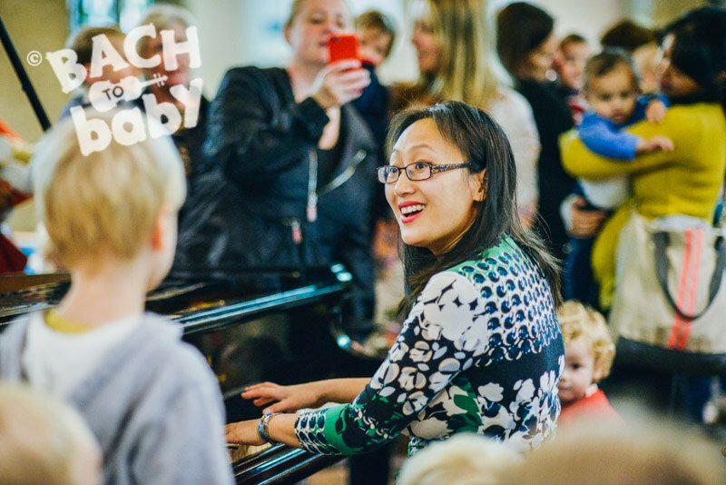 A pianist playing a grand piano, looking to her left and smiling at a boy who is watching her nearby. An out of focus audience of parents/carers and young children in the background