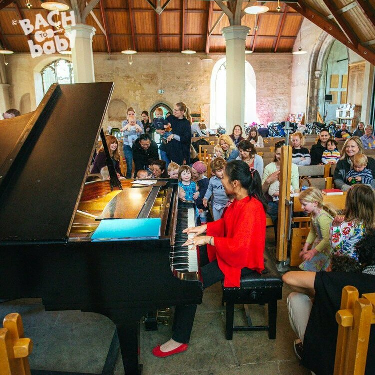 A pianist wearing a red top playing a black grand piano. There's a large audience of young children and parents/carers behind her smiling, and she's turned towards them